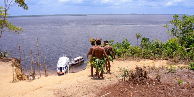Dois homens indígenas observam barcos no rio Amazonas.