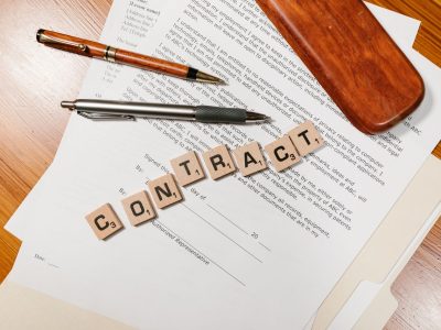 Documentos contratuais sobre uma mesa, com peças formando a palavra “CONTRACT” e uma caneta ao lado