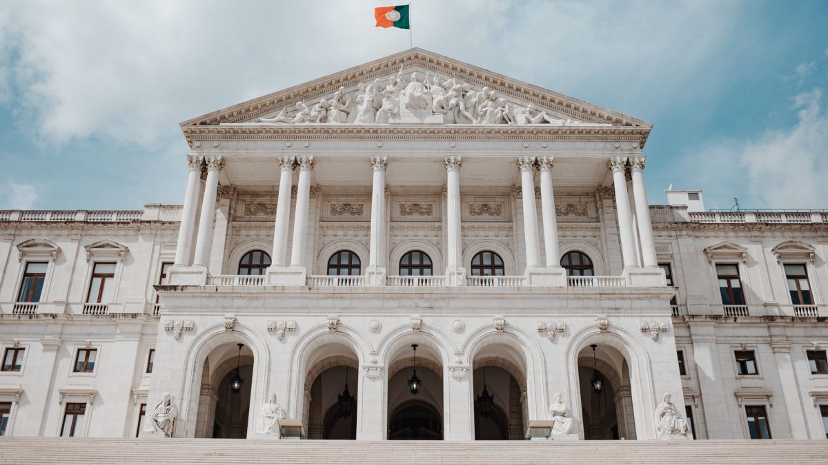Vista frontal do Palácio de São Bento, sede do Parlamento português, com a bandeira nacional hasteada.