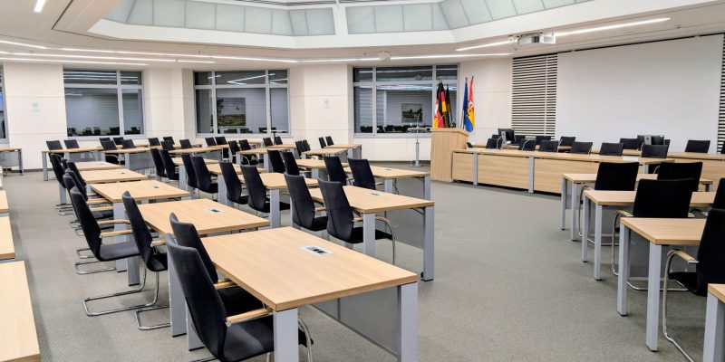 Sala ampla e moderna de reunião institucional, com mesas organizadas e bandeiras ao fundo.
